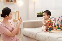 Smiling young mother holding smartphone and photographing adorable baby playing on couch — Stock Photo