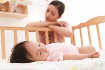 Nahaufnahme des entzückenden Säuglings, der im Kinderbett schläft, und der müden Mutter, die hinten sitzt — Stockfoto