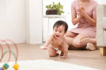 Очаровательный счастливый младенец в подгузнике ползает по полу в то время как мать сидит позади — стоковое фото