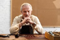 Serious mature man in apron sitting at table with tools and looking at camera at workplace — Stock Photo