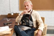 Serious mature man in apron sitting at table with tools and looking at camera at workplace — Stock Photo