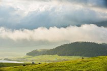 Удивительный горный ландшафт с бунгало на зеленом склоне, водоем и лес в облачный день — стоковое фото