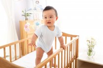 Милий азіатський хлопчик стоїть на дитячому ліжечку і дивиться геть — стокове фото