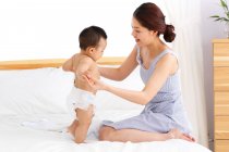 Vue latérale de la jeune mère asiatique jouant avec adorable petit bébé sur le lit — Photo de stock