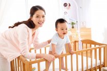 Happy young mother playing with adorable little baby standing in crib — Stock Photo
