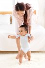 Feliz joven madre aprendiendo adorable pequeño bebé a caminar en casa - foto de stock