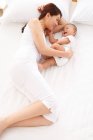Alto ángulo vista de feliz joven madre jugando con adorable pequeño bebé acostado en la cama - foto de stock