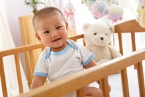 Adorable happy baby boy with teddy bear in crib — Stock Photo