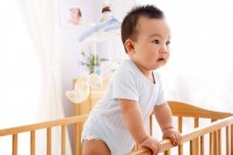 Чарівна допитлива китайська дитина стоїть в ліжечку і дивиться в сторону — стокове фото