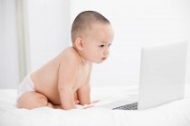 Adorable bebé en pañal sentado en la cama y mirando el ordenador portátil - foto de stock