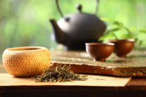 Vista ravvicinata delle foglie di tè essiccate con contenitore e teiera con tazze di ceramica, messa a fuoco selettiva — Foto stock
