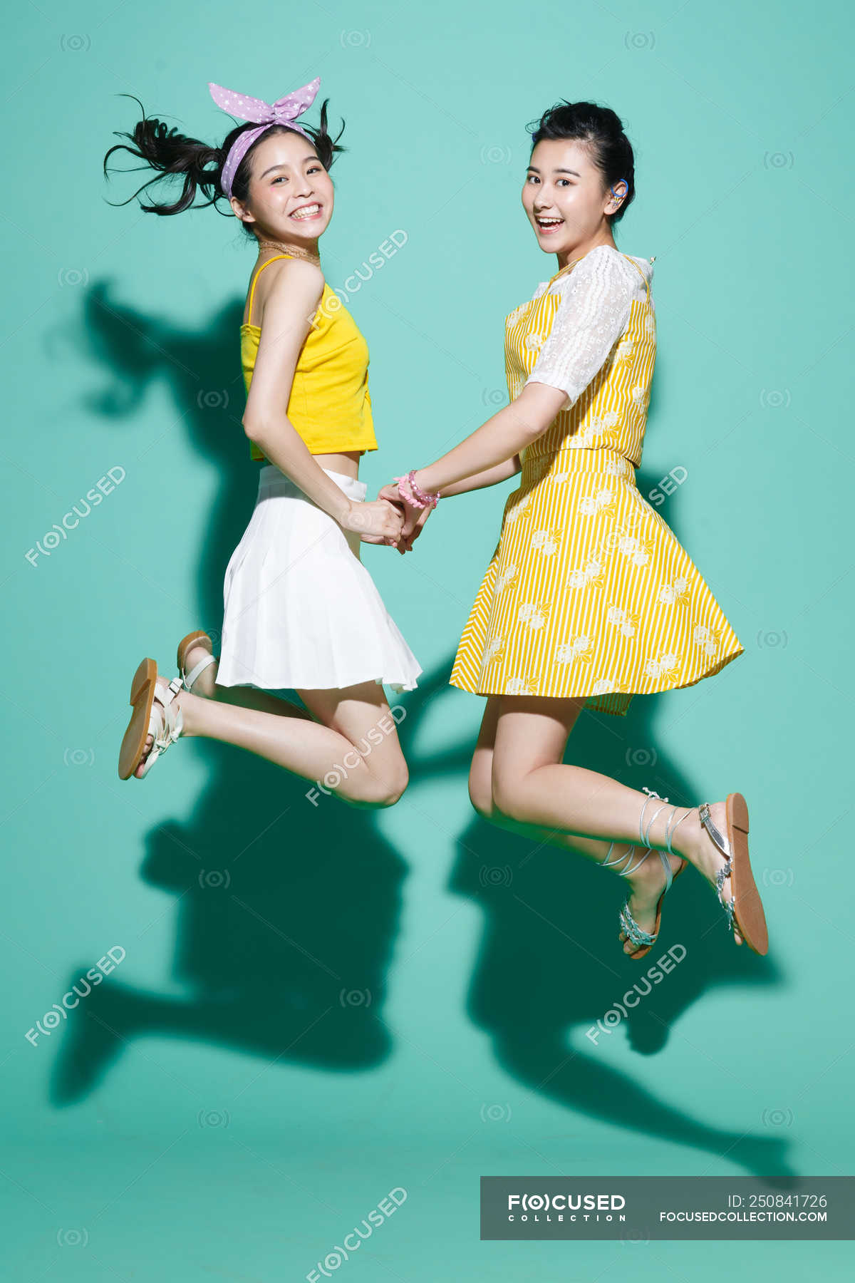 Side View Of Happy Asian Girlfriends Holding Hands And Jumping On Blue — Elegant Models Stock