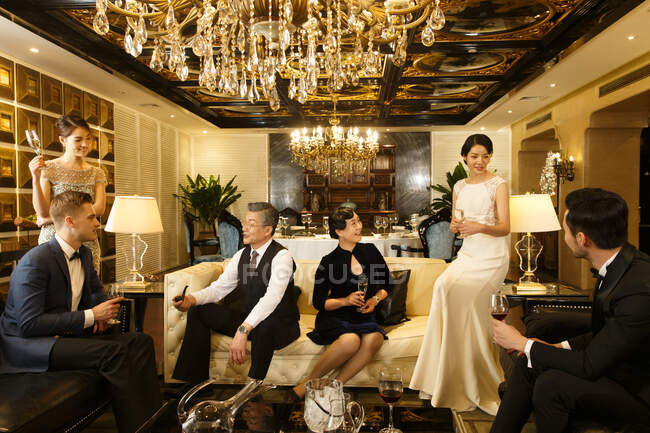 Personas multiétnicas elegantes en trajes y vestidos de noche beber champán, pipa de fumar e interactuar en la habitación de lujo - foto de stock