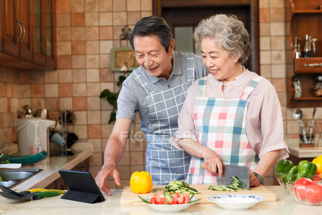 Пожилая азиатская пара с помощью цифрового планшета и приготовления пищи вместе дома — стоковое фото