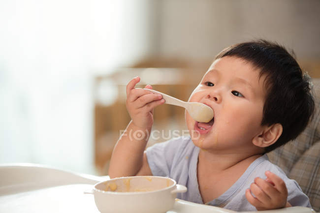 Adorable asian toddler holding spoon and eating at home — Stock Photo