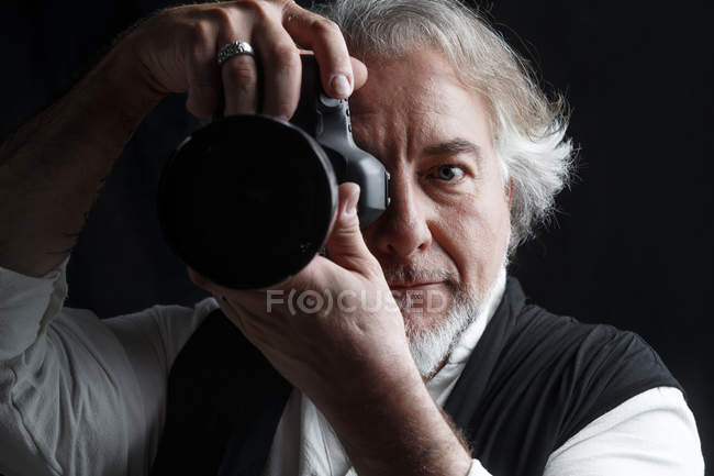 Fotógrafo maduro profesional utilizando la cámara de fotos y mirando la cámara aislada en negro - foto de stock
