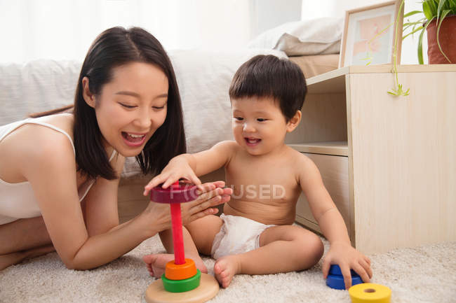 Feliz jovem mãe batendo palmas e olhando para o bebê sorrindo brincando com brinquedo colorido em casa — Fotografia de Stock