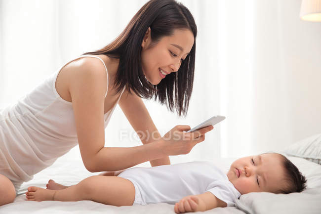 Улыбающаяся молодая азиатская женщина с смартфоном и фотографирующая очаровательного ребенка спящего на кровати — стоковое фото