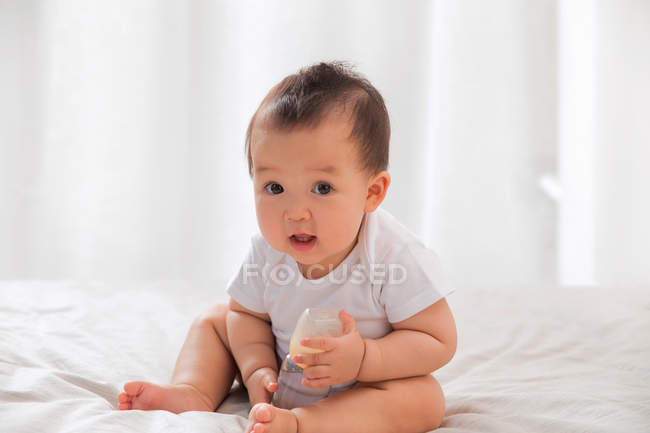 Lovely asian baby sitting on bed wity baby bottle and looking at camera — Stock Photo