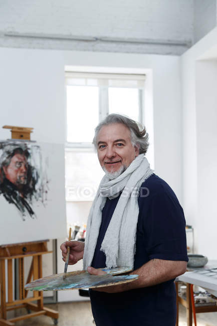 Happy mature male artist holding palette with brush and smiling at camera in studio — Stock Photo