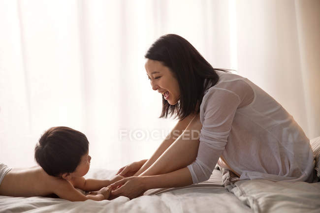 Vista lateral de la feliz madre joven sentada en la cama y jugando con un niño adorable - foto de stock