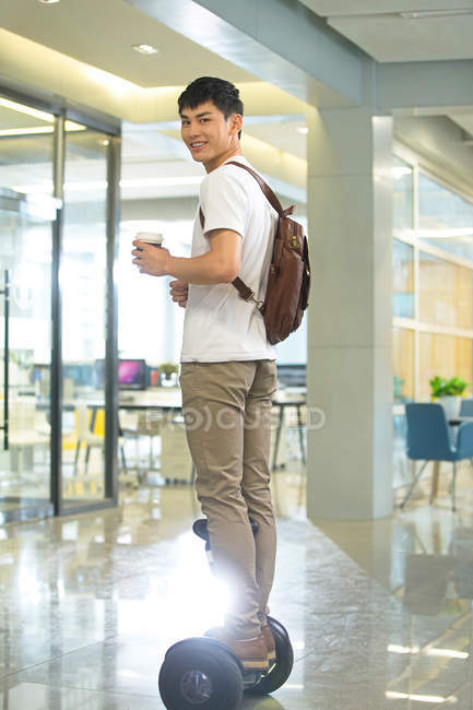 Giovane uomo d'affari sorridente con zaino che tiene il caffè per andare e cavalcare scooter auto-bilanciamento in ufficio — Foto stock
