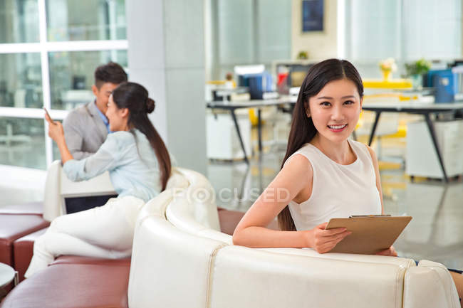 Bella giovane donna d'affari asiatica che tiene appunti e sorride alla fotocamera in ufficio — Foto stock