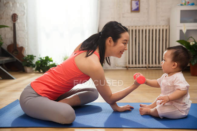 Вид сбоку на молодую маму, которая занимается с гантелями и смотрит на симпатичного ребенка, сидящего дома на коврике для йоги — стоковое фото