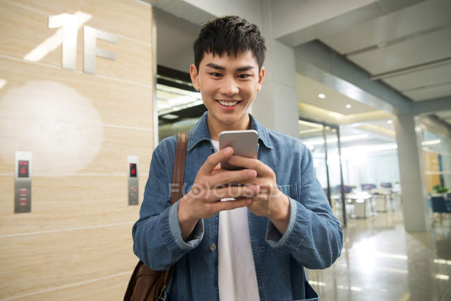 Усміхнений молодий азіатський чоловік використовує смартфон в офісі — стокове фото