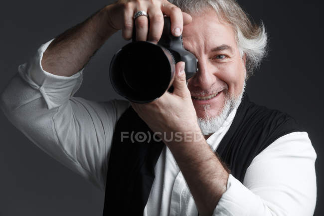 Профессиональный мужчина-фотограф, работающий с фотокамерой и улыбающийся на камеру, изолированную на сером — стоковое фото
