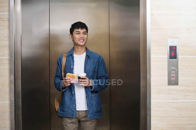 Lächelnder junger asiatischer Geschäftsmann mit Brötchen und Kaffee, um im Fahrstuhl zu stehen und wegzuschauen — Stockfoto