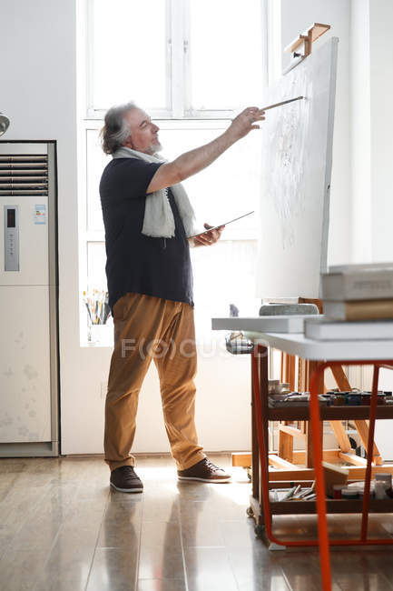 Side view of male artist holding palette with brush and painting in studio — Stock Photo
