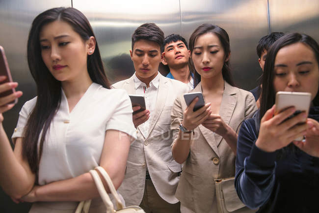 Серьезные молодые азиатские люди, использующие смартфоны в лифте — стоковое фото