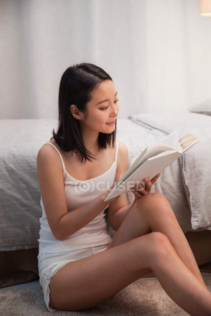 Schöne lächelnde junge asiatische Frau sitzt in der Nähe des Bettes und liest Buch — Stockfoto