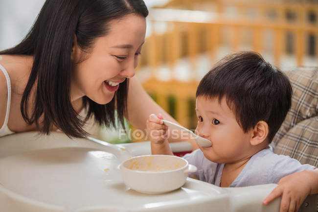 Happy young mother looking at adorable little child eating at home — Stock Photo