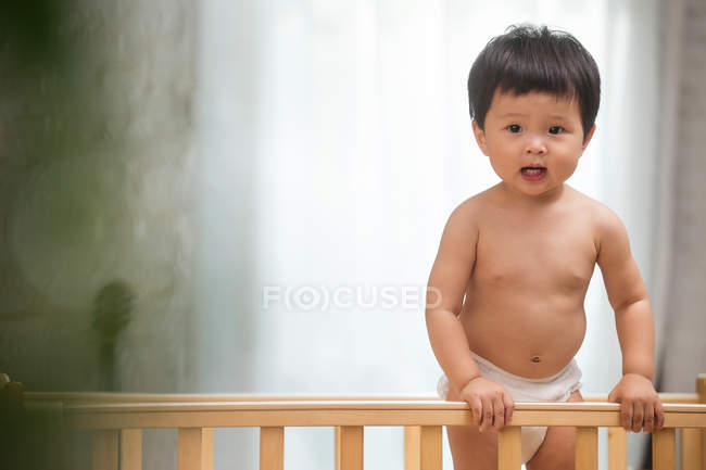Adorable asian toddler in diaper standing in crib and looking at camera — Stock Photo