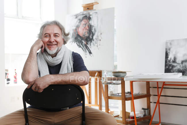 Красивый счастливый зрелый художник, сидящий на стуле и улыбающийся камере в студии — стоковое фото