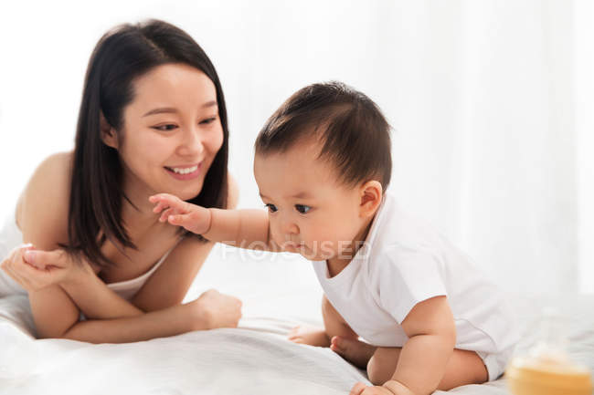 Красивая счастливая молодая азиатская мать смотрит на своего очаровательного ребенка, сидящего на кровати — стоковое фото