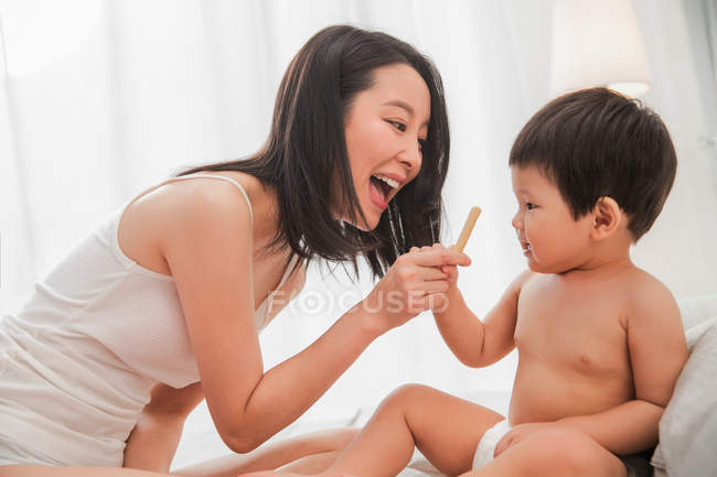 Vista lateral de feliz joven madre sosteniendo palo y jugando con su precioso bebé en casa - foto de stock