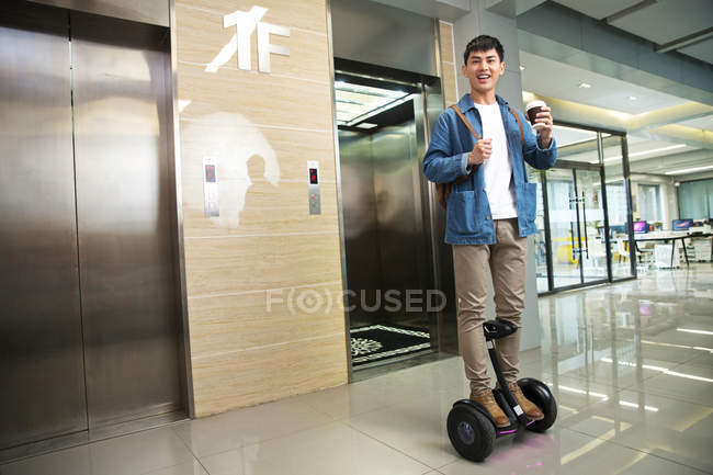 Handsome young asian businessman on self-balancing scooter holding coffee to go near elevator — Stock Photo