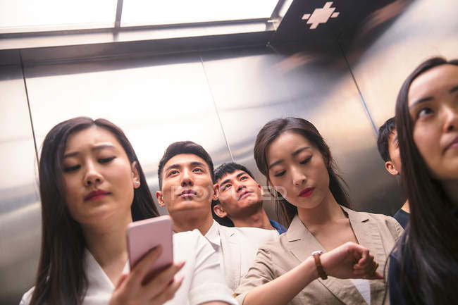 Bajo ángulo vista de serios jóvenes asiáticos hombres de negocios y mujeres de negocios de pie juntos en ascensor - foto de stock