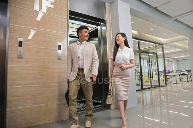 Young asian businessman and businesswoman smiling each other while walking from elevator in office — Stock Photo