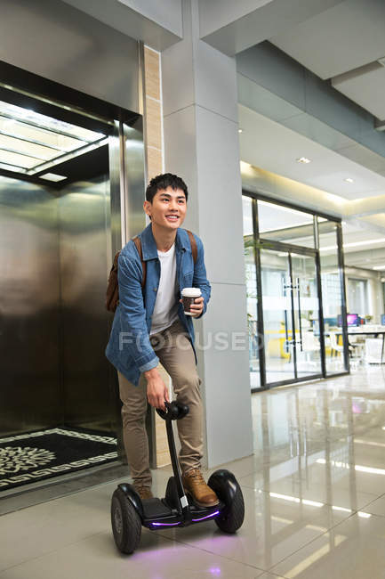 Handsome young asian businessman on self-balancing scooter holding coffee to go near elevator — Stock Photo