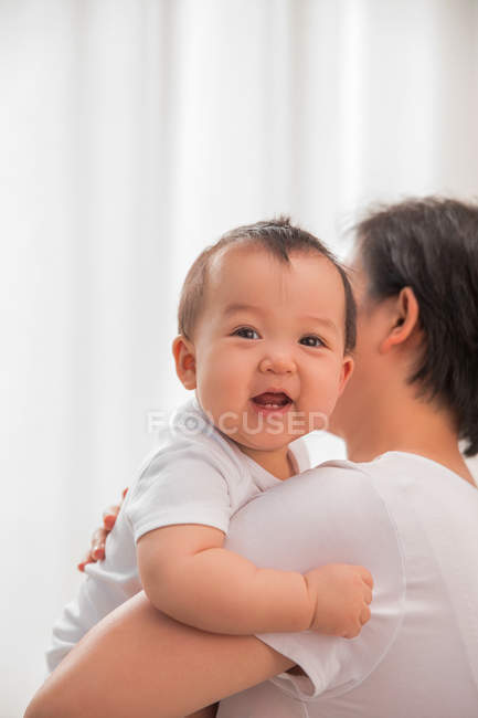 Мати носить чарівну азіатську дитину, посміхаючись на камеру вдома — стокове фото