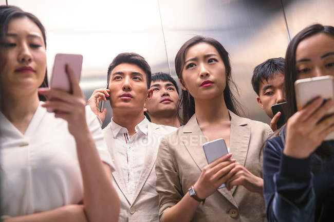 Серьезные молодые азиатские люди, использующие смартфоны в лифте — стоковое фото