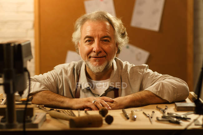 Professionelle reife männliche Schmuckdesignerin sitzt am Arbeitsplatz und lächelt in die Kamera — Stockfoto