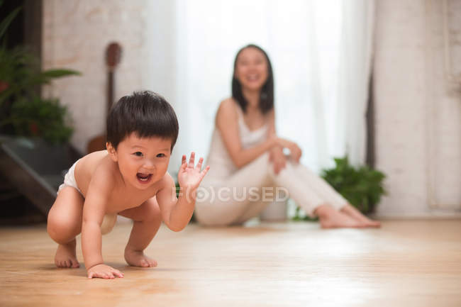 Очаровательный взволнованный азиатский ребенок в подгузнике, приседающий на полу, в то время как счастливая мать сидит позади, избирательный фокус — стоковое фото