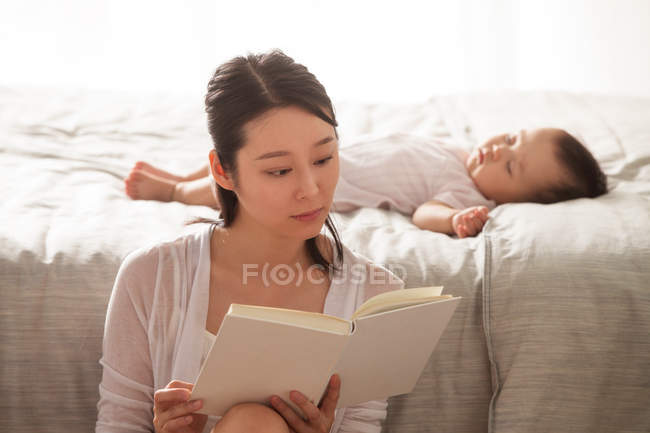 Сосредоточена молодая азиатская женщина читает книгу, пока ребенок спит на кровати позади — стоковое фото