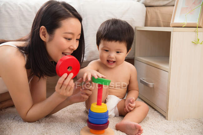 Счастливая молодая мама помогает восхитительной улыбчивой малышке играть с цветной игрушкой дома — стоковое фото