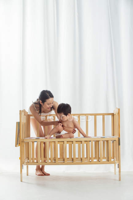 Volle Länge Ansicht der glücklichen jungen Mutter spielt mit entzückenden Kleinkind in Windel sitzen in der Krippe — Stockfoto